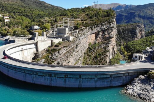 Vue aérienne du barrage TDAMB entouré de montagnes et d'eau turquoise.