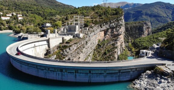 Vue aérienne du barrage TDAMB entouré de montagnes et d'eau turquoise.